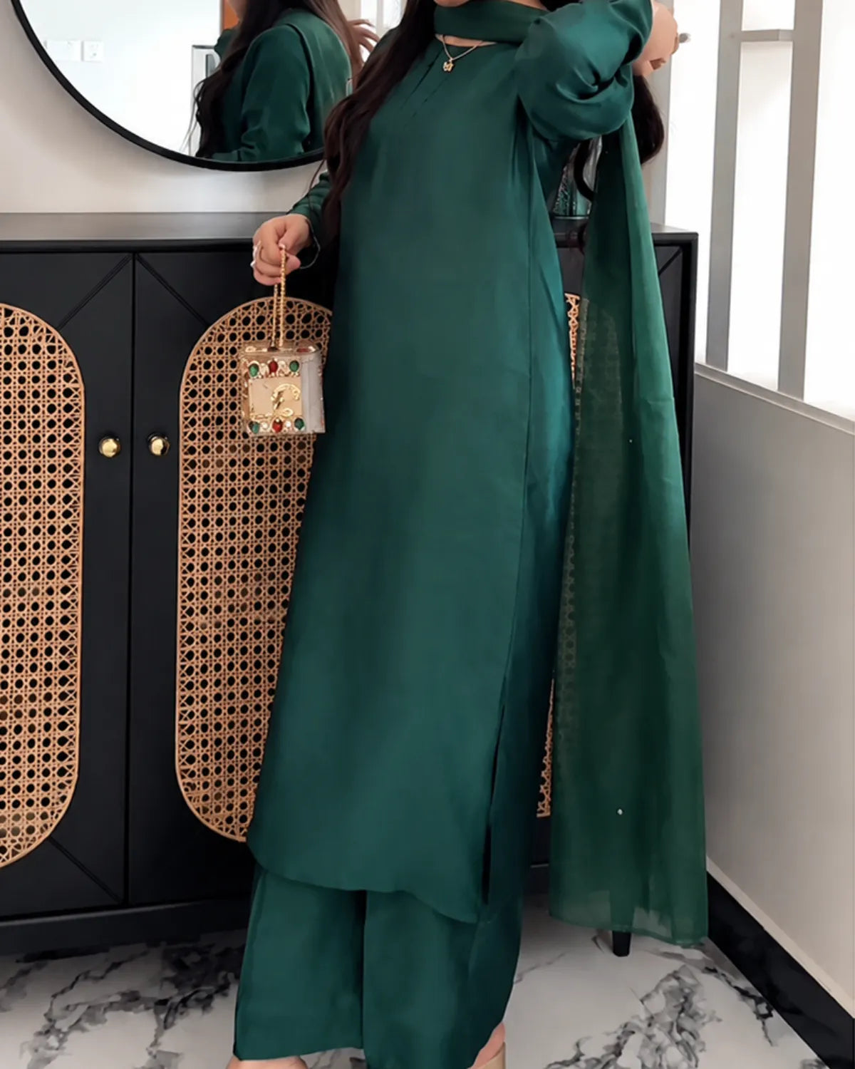 A woman models a dark green ethnic outfit consisting of a long-sleeve kurta, matching wide-leg pants, and a sheer dupatta, standing in front of a black cabinet with woven panels.