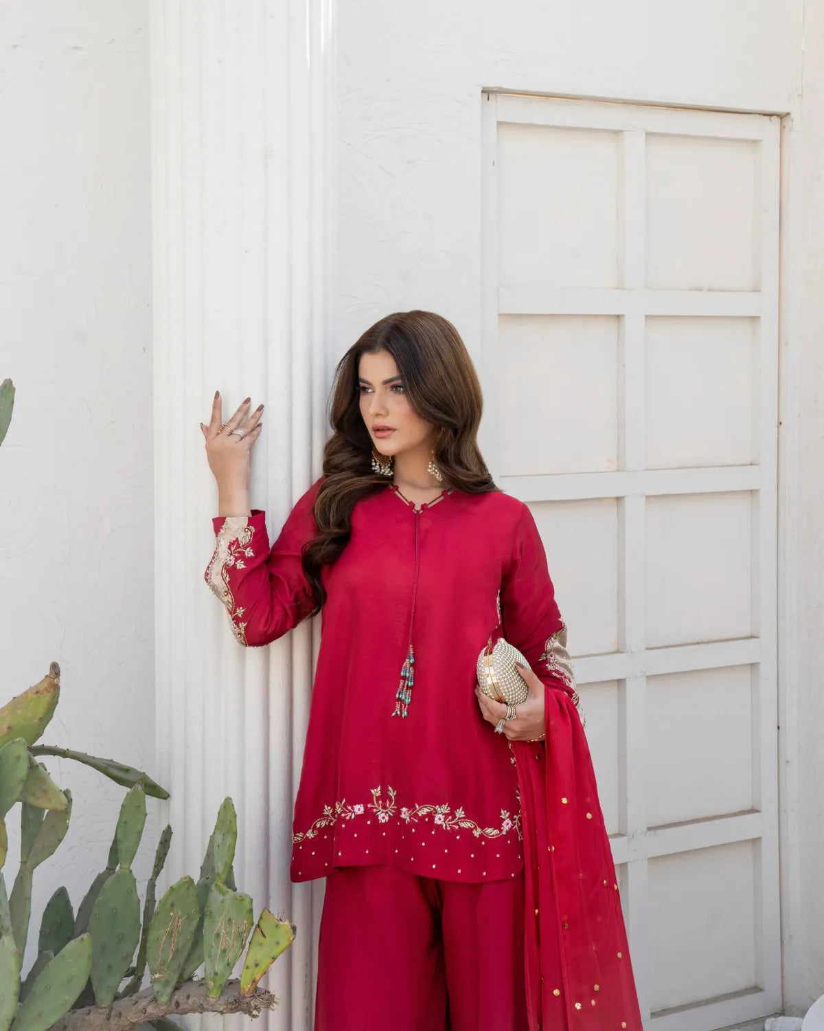 A woman models a bright red three-piece ethnic suit with floral embroidery, featuring a kurta, matching trousers, and a dupatta, posed against a white stucco wall.