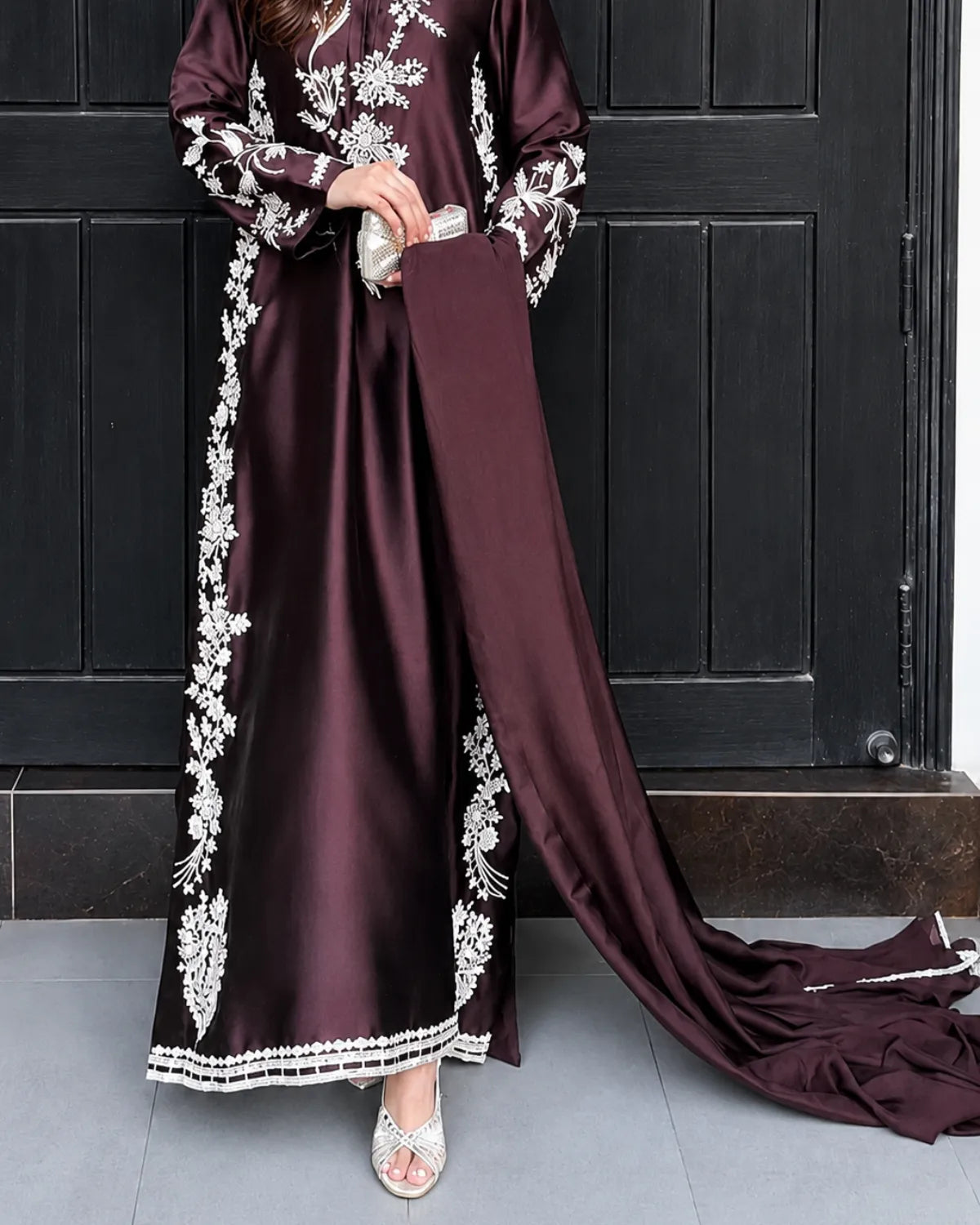 A woman wearing a long, floor-length maroon satin gown with white floral embroidery and a matching draped train, standing in front of a dark paneled door.