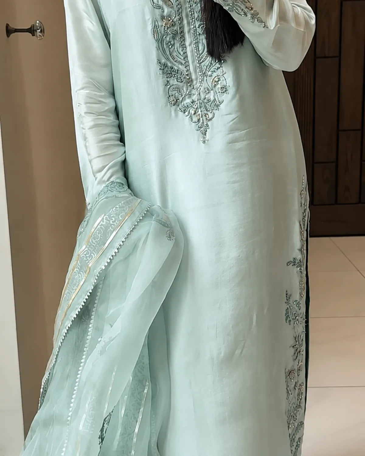 A person wearing a light mint green traditional outfit with silver embroidery on the chest and a matching sheer dupatta draped over one arm, shown indoors.