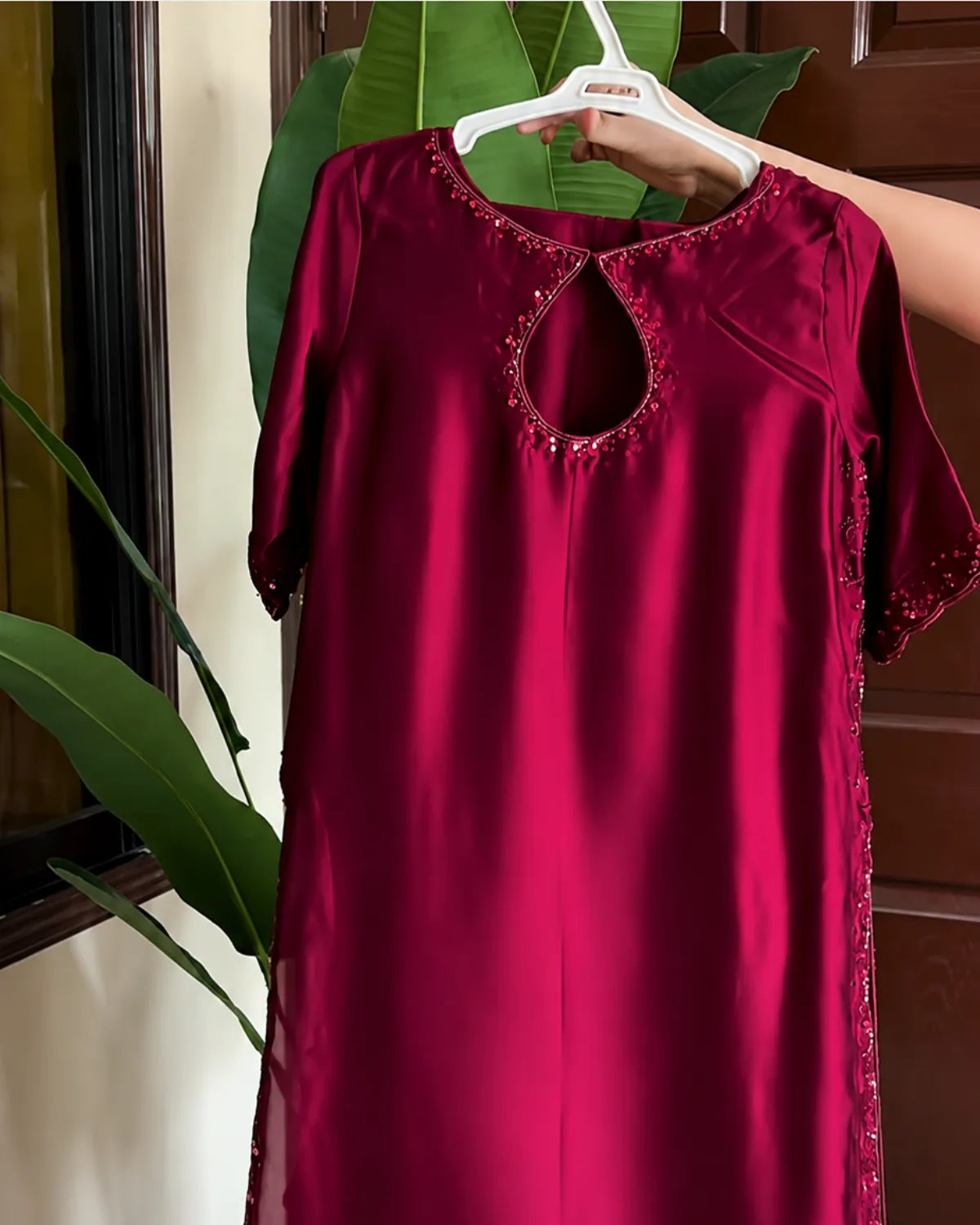 A deep magenta-red satin nightgown with a keyhole cutout and embroidered details, held up on a white hanger by a hand in front of a background of green leaves and a wooden door.