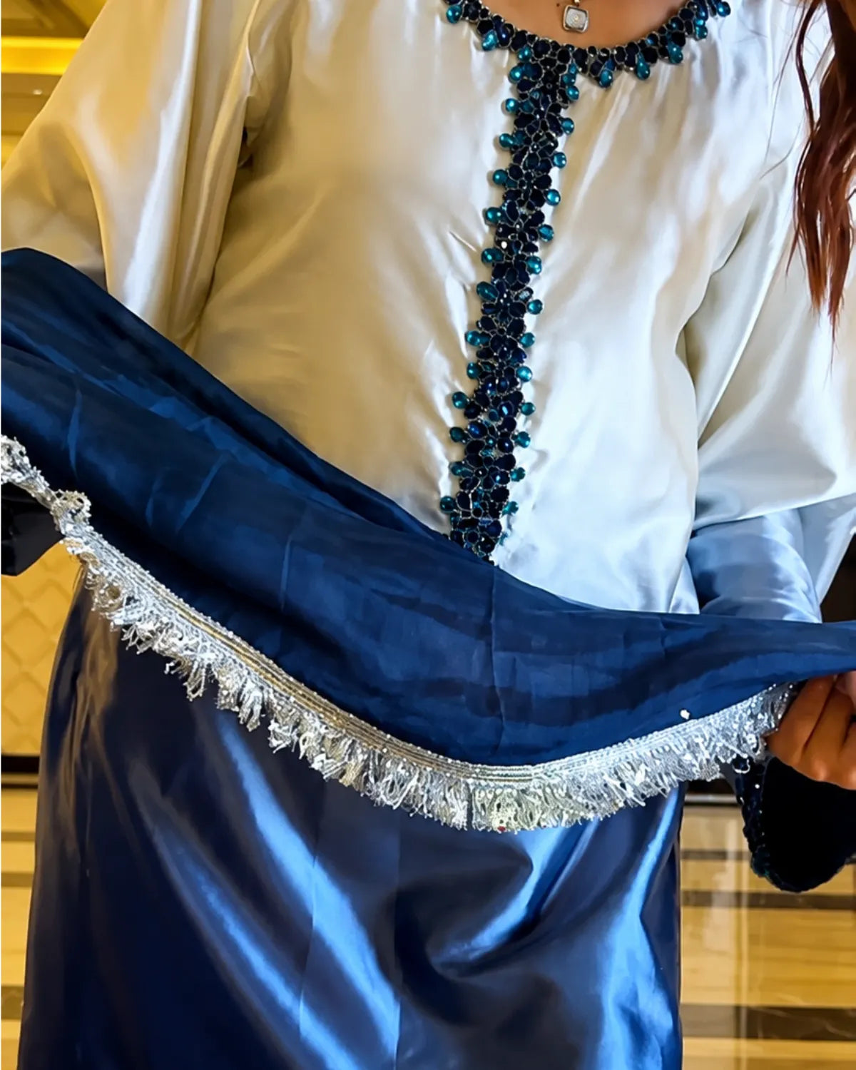 A close-up of a person wearing a white satin-like top with a dark blue beaded neckline and holding a dark blue dupatta with a silver embroidered fringe.