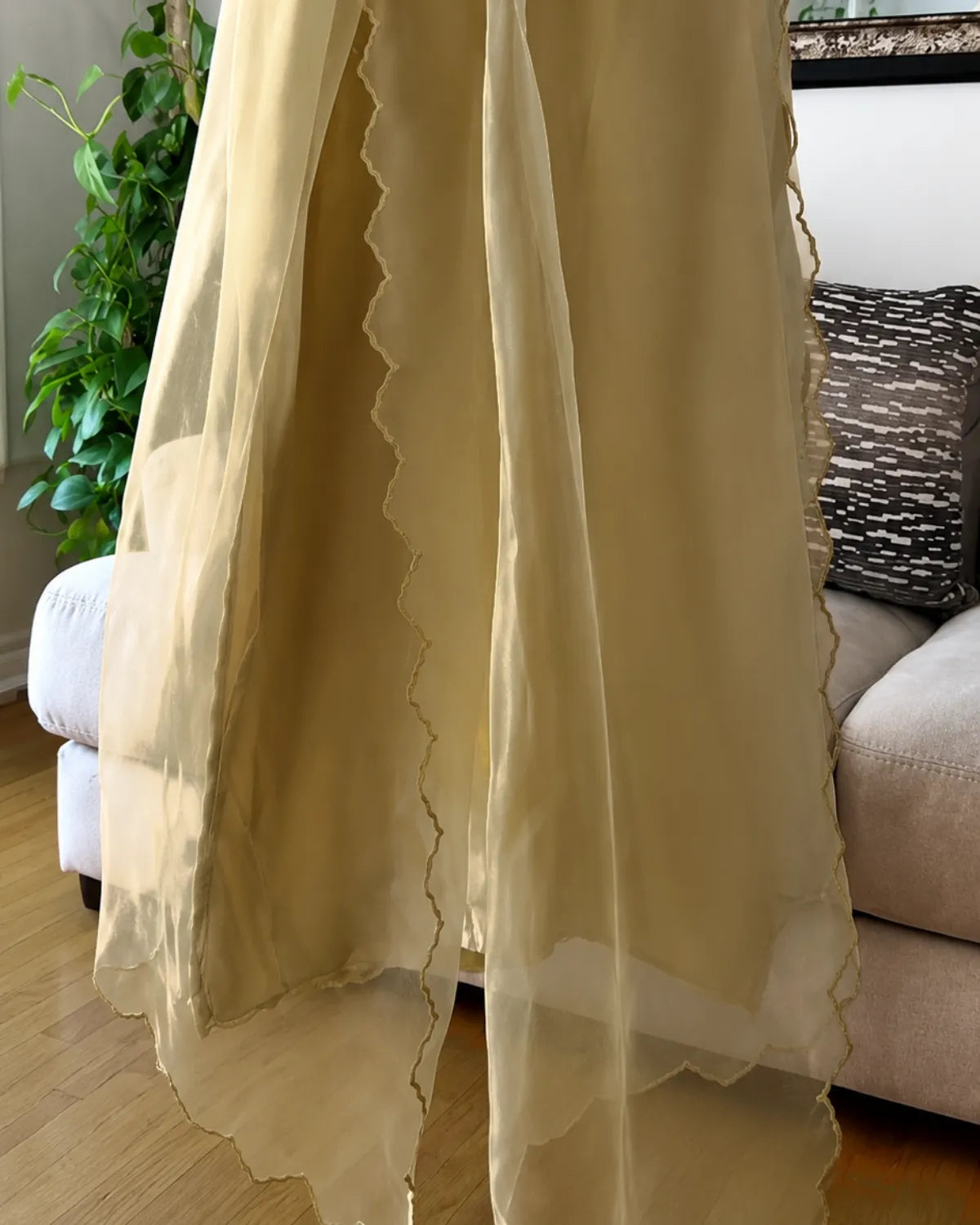 A sheer beige curtain panel with scalloped edges hanging in front of a window in a living room setting.