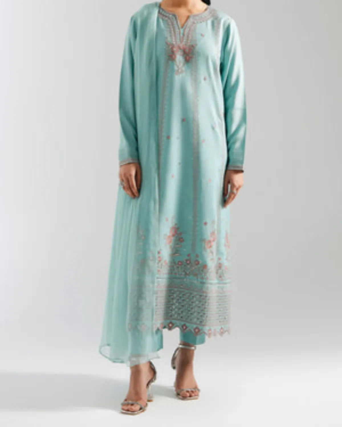 A woman modeling a long mint green kurta with pink and gold embroidery, paired with matching pants and heeled sandals, against a light gray studio background.