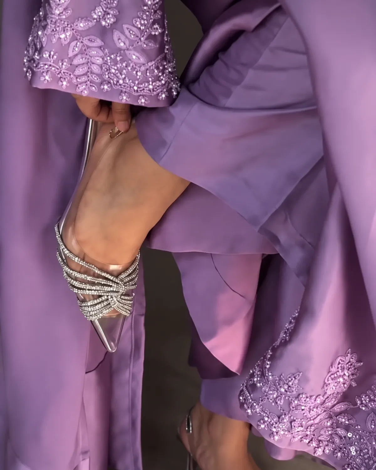 A close-up lifestyle shot of a person wearing a flowing lavender dress with intricate silver beadwork and embellishments, adjusting a clear high-heeled shoe with multiple silver rhinestone straps.