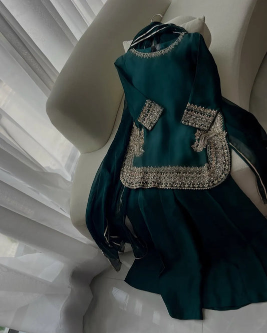 A dark teal green traditional outfit with intricate silver embroidery, draped on a white chair in front of sheer white curtains.