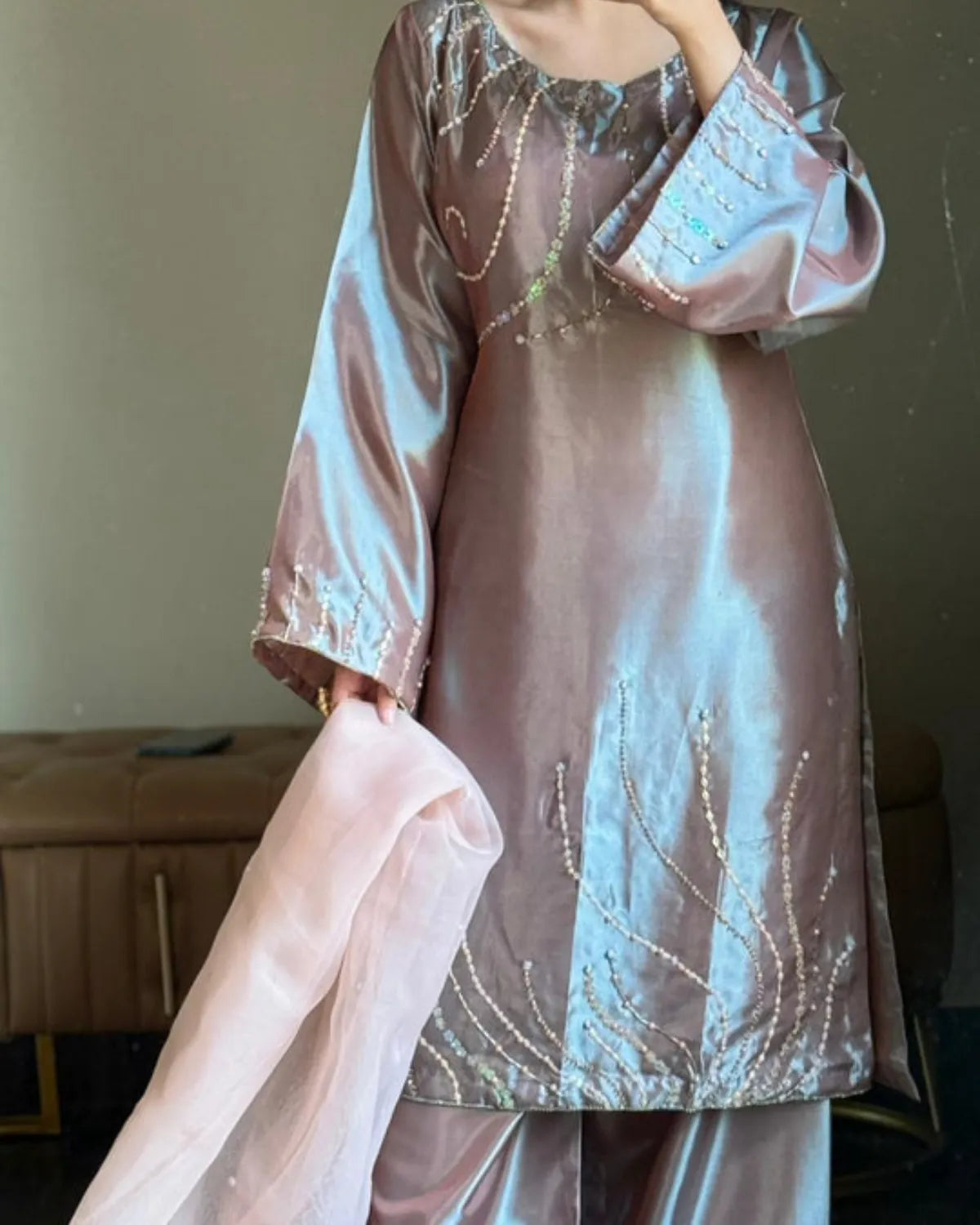 A person wearing a shiny taupe-brown satin kurta and matching trousers, holding a light pink dupatta. The kurta features silver beadwork embroidery and has a round neckline, photographed in an indoor setting.