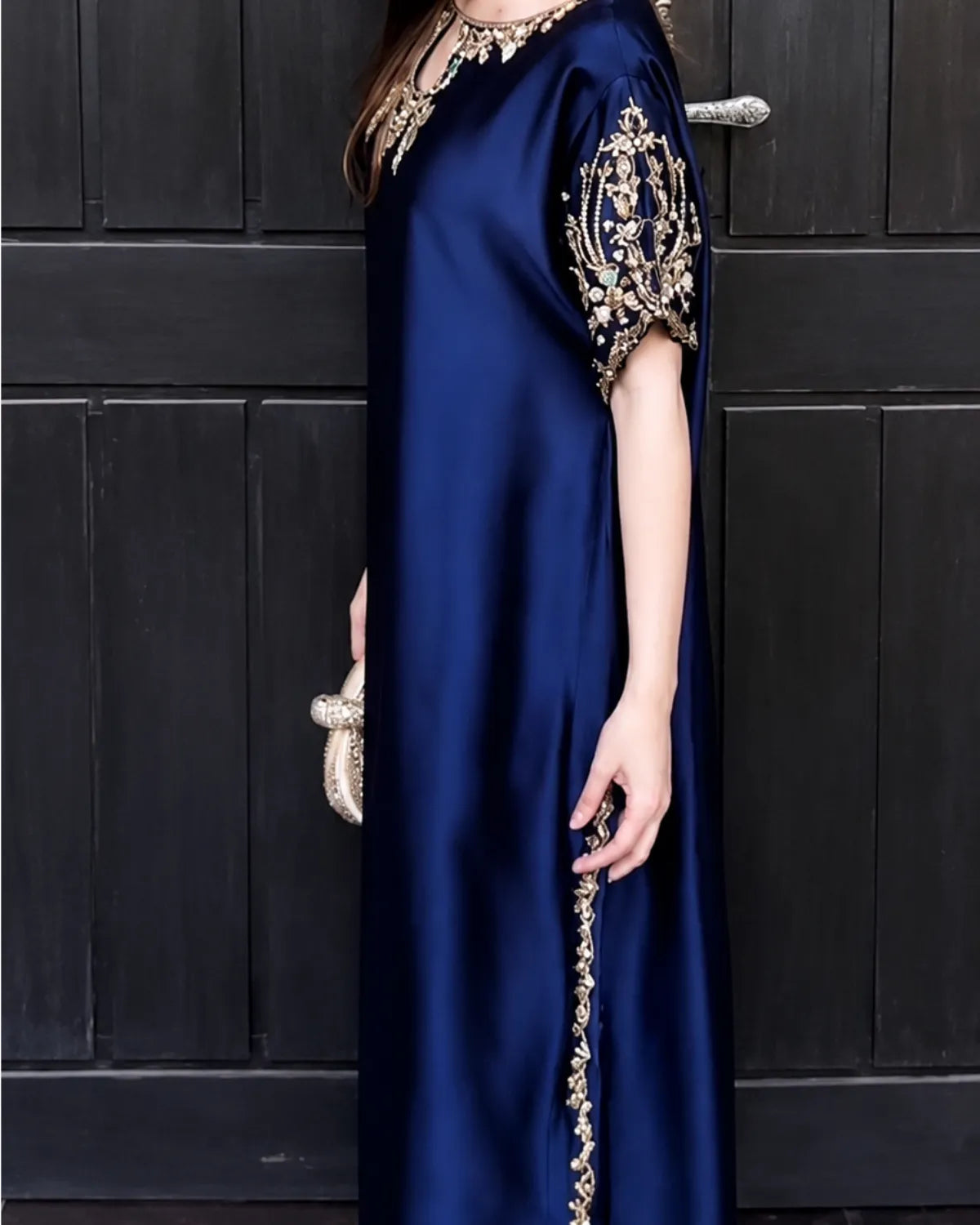 A woman wearing a navy blue satin maxi dress with ornate gold embroidery on the sleeve and neckline, standing in front of a dark paneled door.