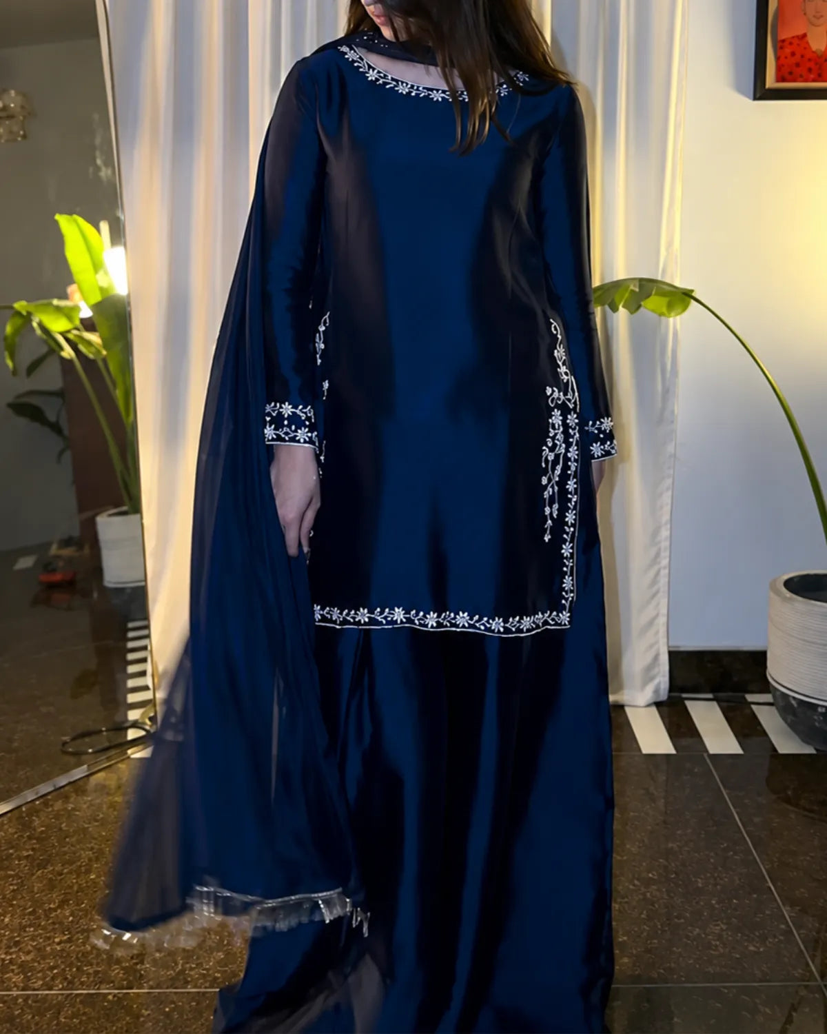 A woman wearing a navy blue traditional outfit with white embroidered details, standing indoors in front of white curtains and potted plants.