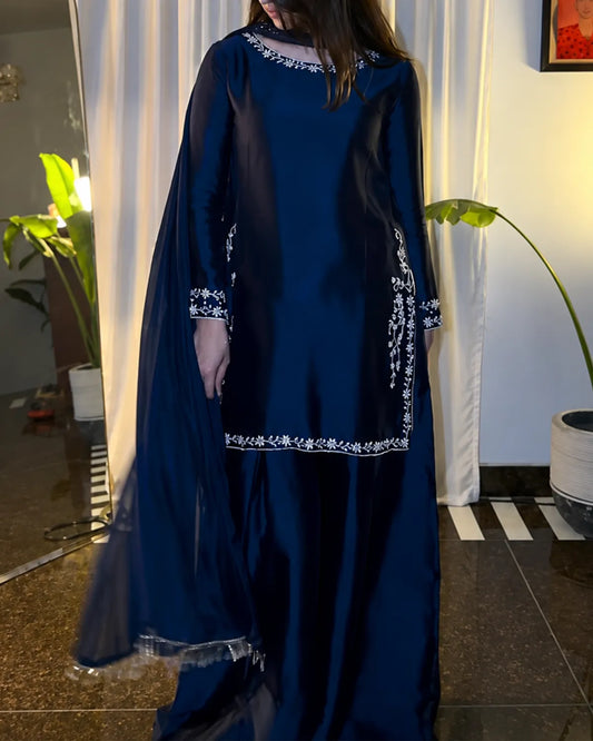A woman wearing a navy blue traditional outfit with white embroidered details, standing indoors in front of white curtains and potted plants.