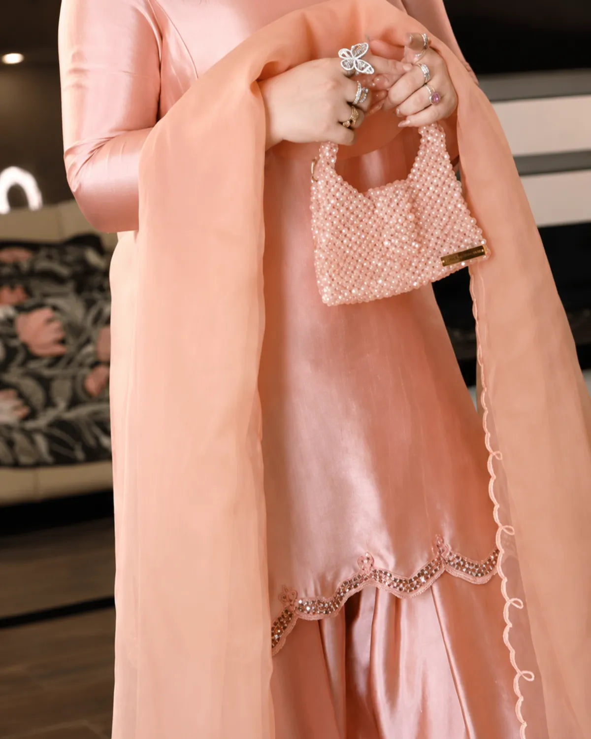 A person wearing a peach-pink satin traditional outfit with a matching sheer dupatta and holding a small embellished peach-pink handbag. The outfit has scalloped embroidered detailing near the hem.