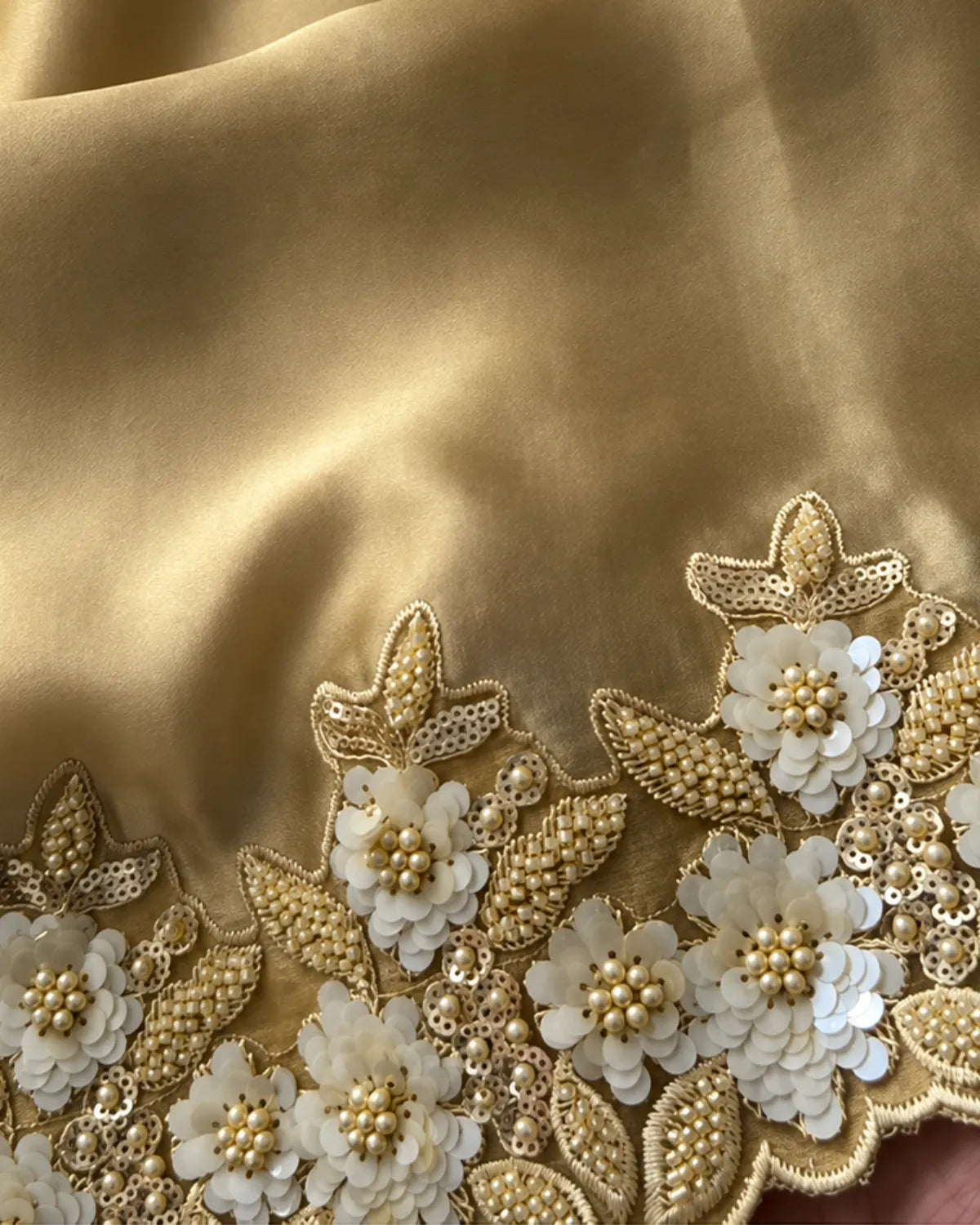 Close-up of a gold satin fabric with intricate floral embroidery and white sequined flowers along the border.