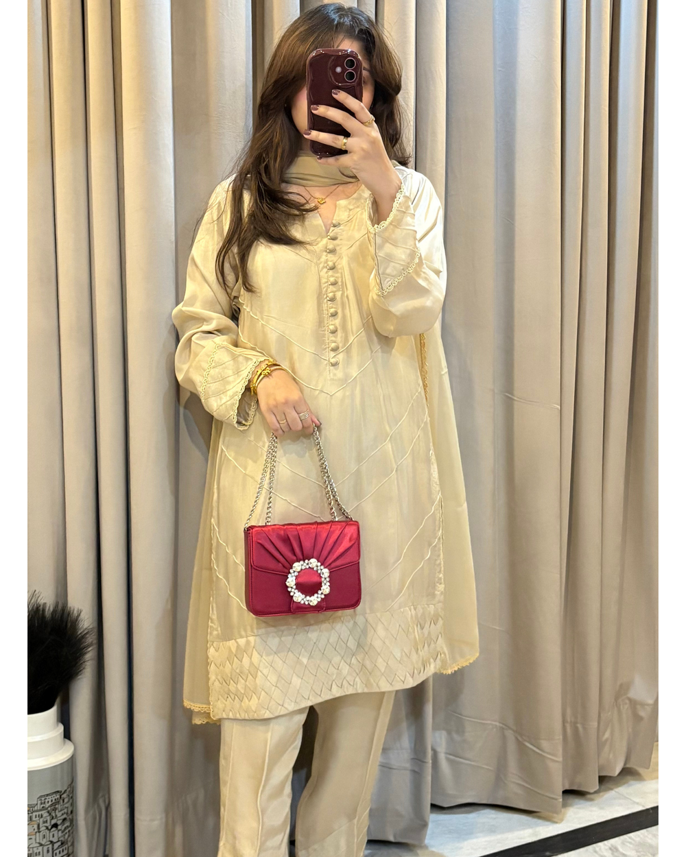 Person wearing a light-colored outfit with a red handbag in front of beige curtains.