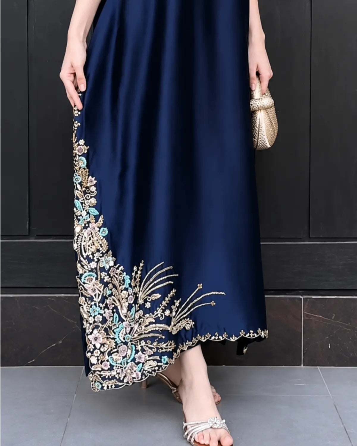 A navy blue maxi dress with elaborate gold, silver, and turquoise beadwork embroidery along the hem and side, shown on a model holding a small gold clutch against a dark paneled wall.