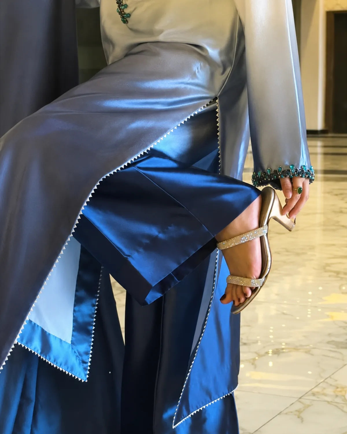A close-up of a person wearing a flowing blue satin garment with layered panels, decorative beaded trim, and heeled sandals in an indoor setting with a marble floor.