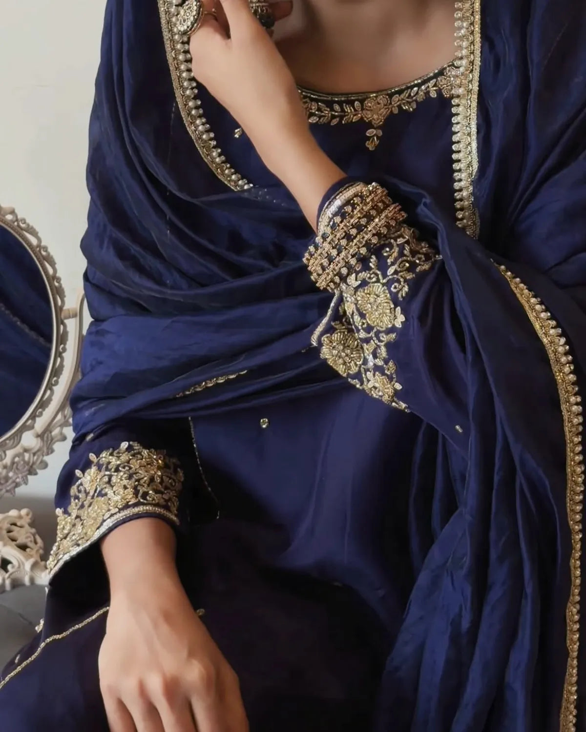 A close-up of a person wearing a deep navy blue traditional outfit with ornate gold embroidery on the neckline, sleeve cuffs, and draped dupatta.