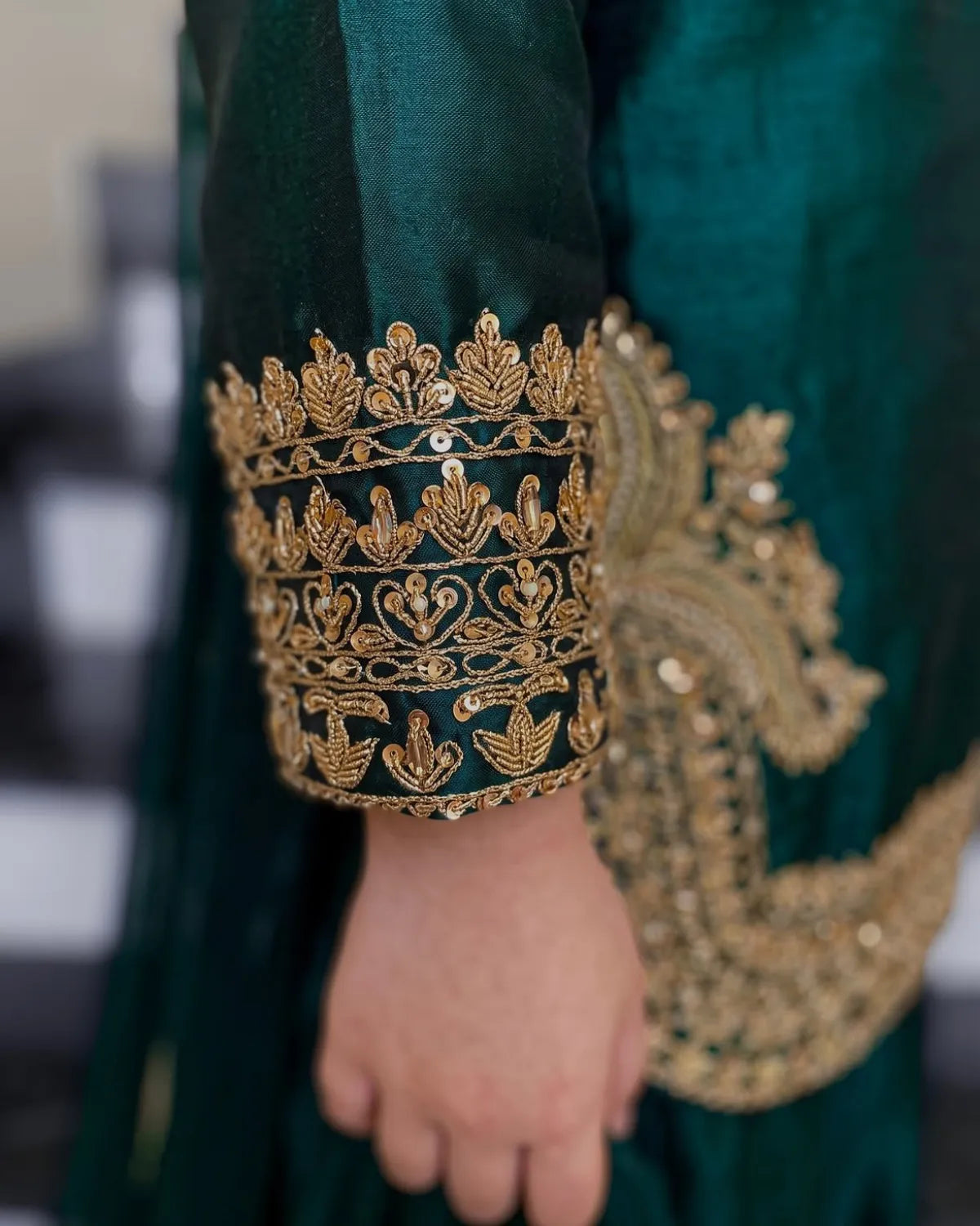 Close-up of a dark teal green traditional garment with ornate gold embroidery on the sleeve cuff and a matching paisley motif on the body, worn by a person whose hand is visible.