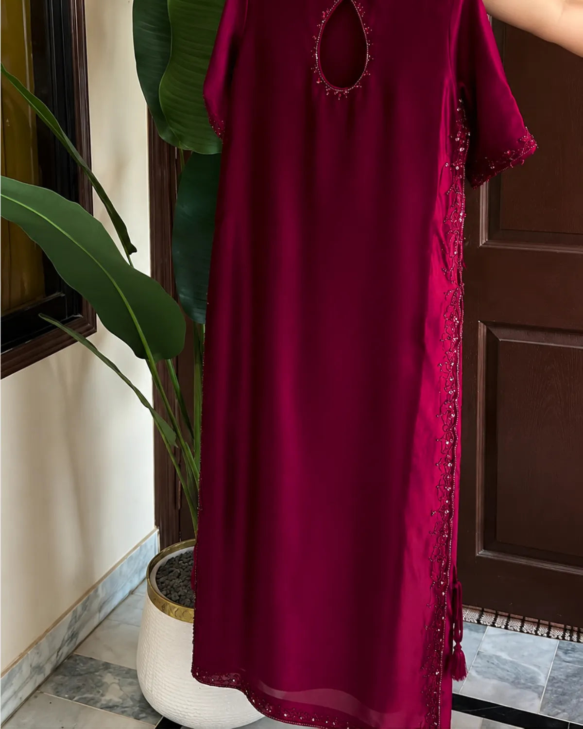A deep magenta-red maxi dress with short sleeves, a keyhole cutout at the neckline, and decorative beadwork along the side, shown hanging indoors next to a potted plant.