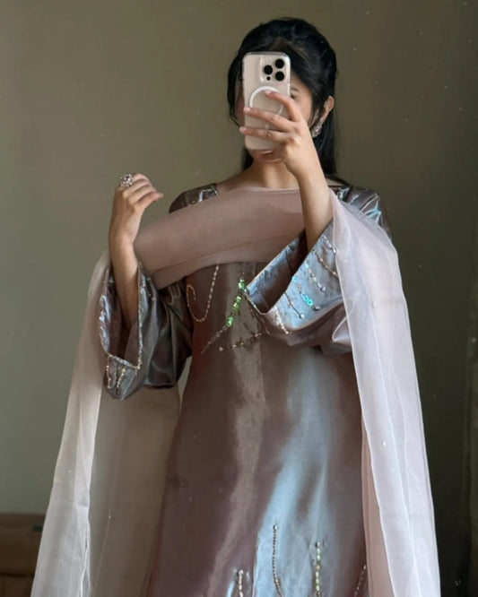 A woman taking a mirror selfie wearing a traditional outfit consisting of a dusty rose dress with metallic silver sleeves and a sheer light pink dupatta.