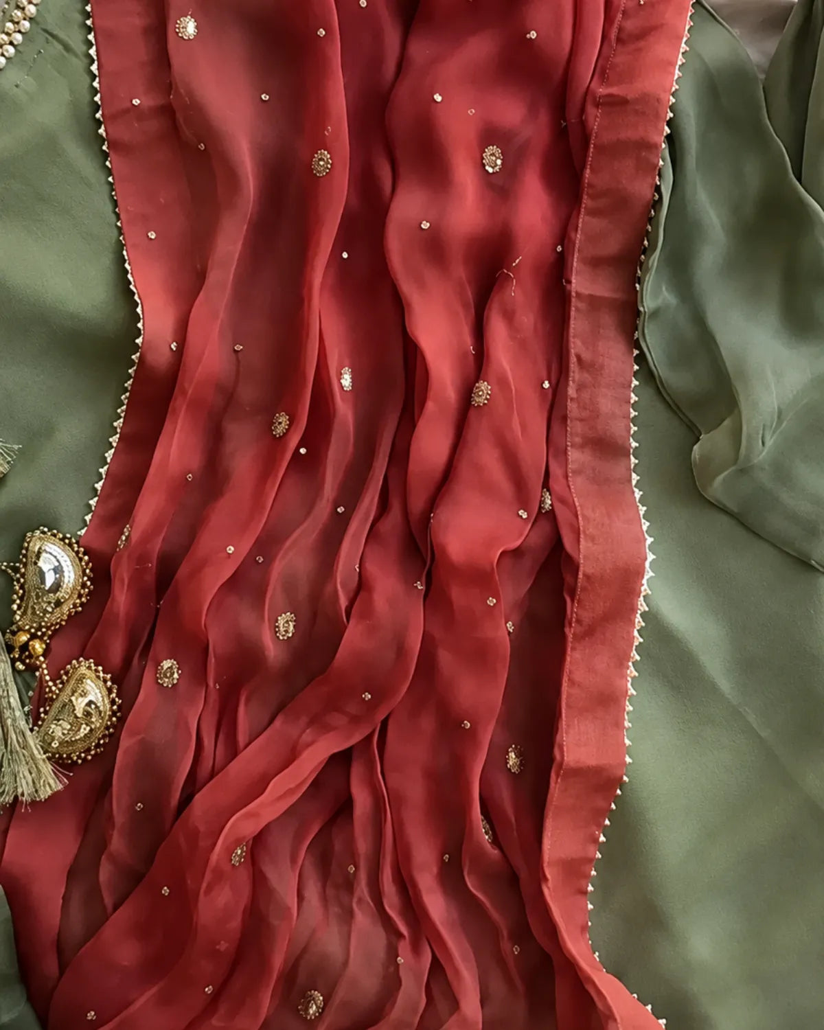 A close-up of a draped terracotta red sheer fabric with gold embroidered motifs and a scalloped edge, laid over an olive green fabric with a decorative gold tassel ornament.