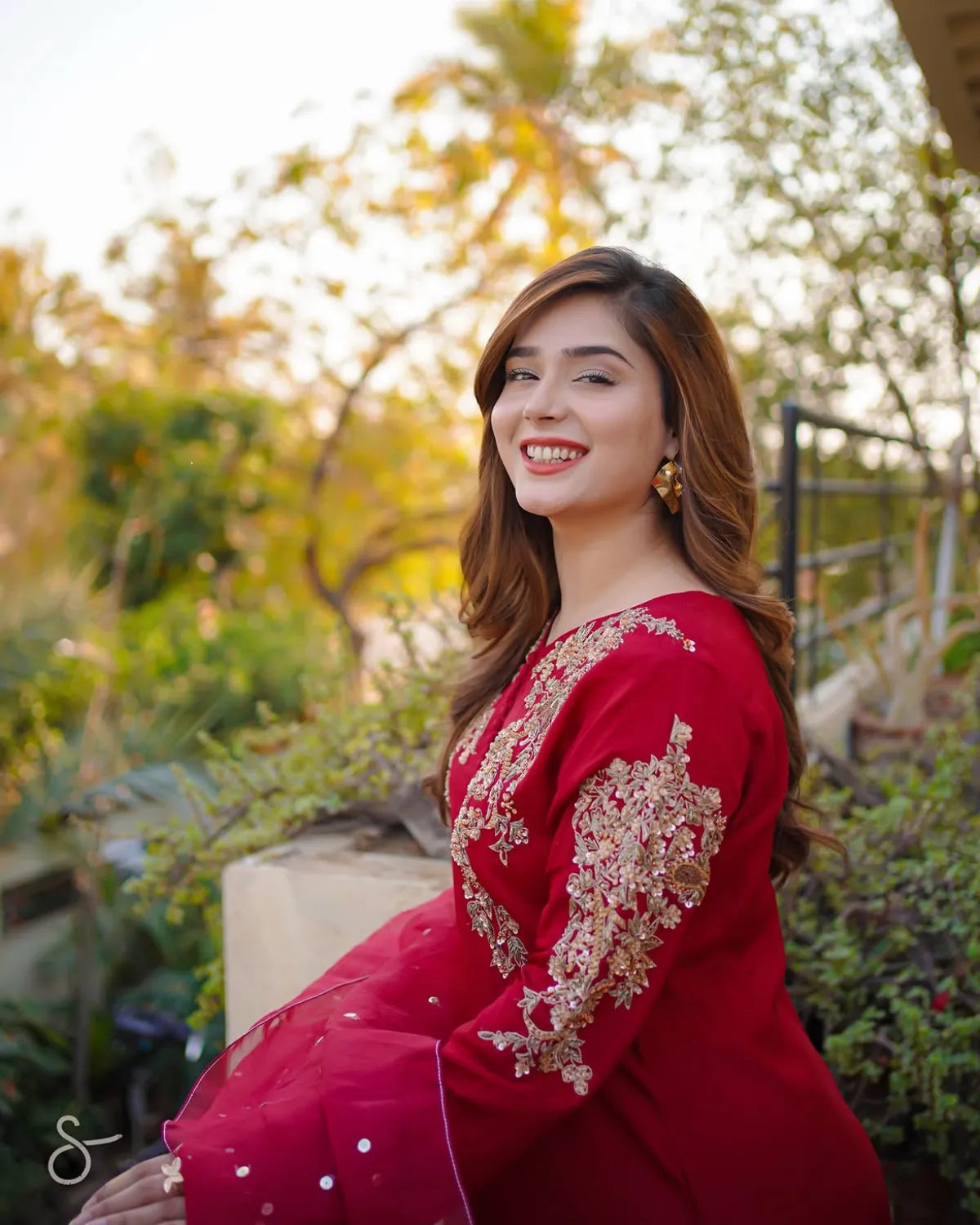 A woman wearing a red traditional embroidered outfit with gold detailing, photographed outdoors in a garden setting with warm sunlight and blurred greenery in the background.