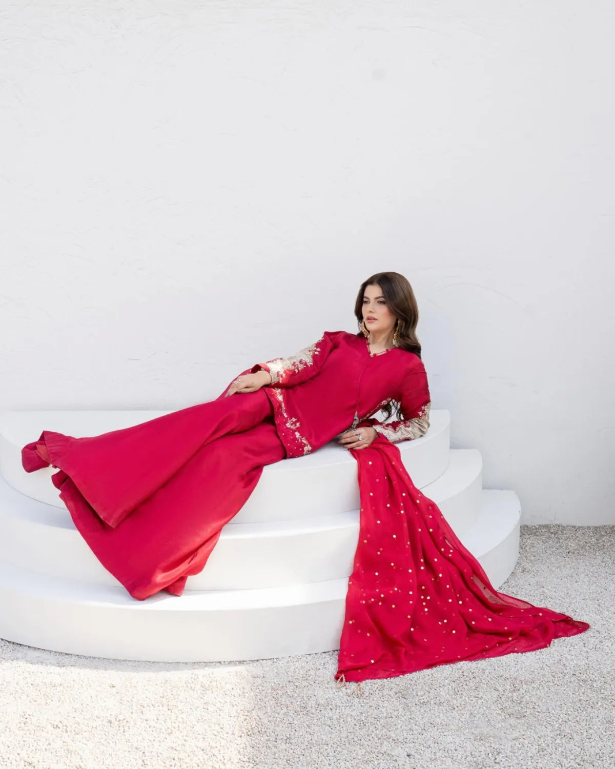 A woman reclines on white tiered steps wearing a bright red traditional outfit with sheer embroidered sleeves and a long draped skirt that pools on the floor against a white wall.
