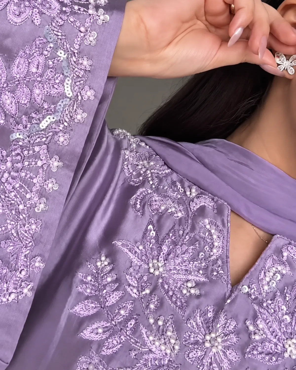 A close-up of a person wearing a lavender satin ethnic outfit with intricate silver and white floral embroidery, beadwork, and a matching draped scarf. The person is shown from the chest up, with one hand near the face, wearing a silver butterfly-shaped earring.