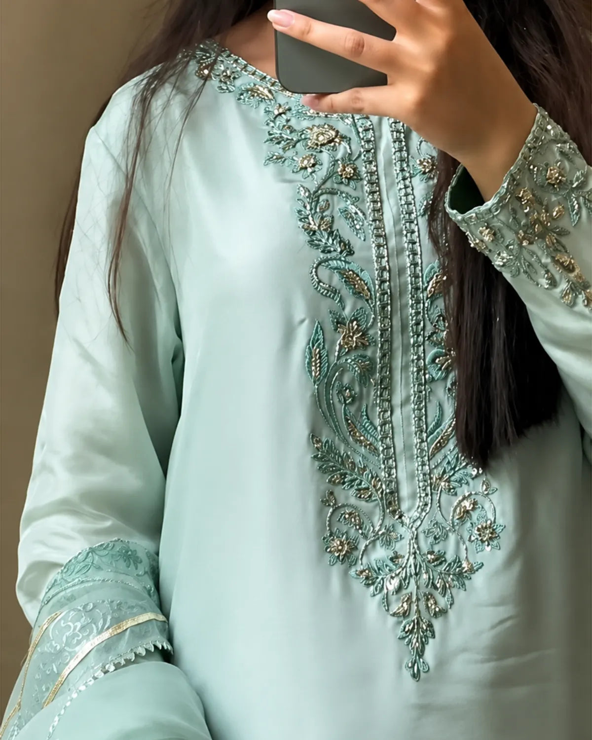 A close-up of a person wearing a mint green traditional embroidered tunic with ornate metallic embroidery on the chest and cuffs, holding a smartphone to take a mirror selfie.