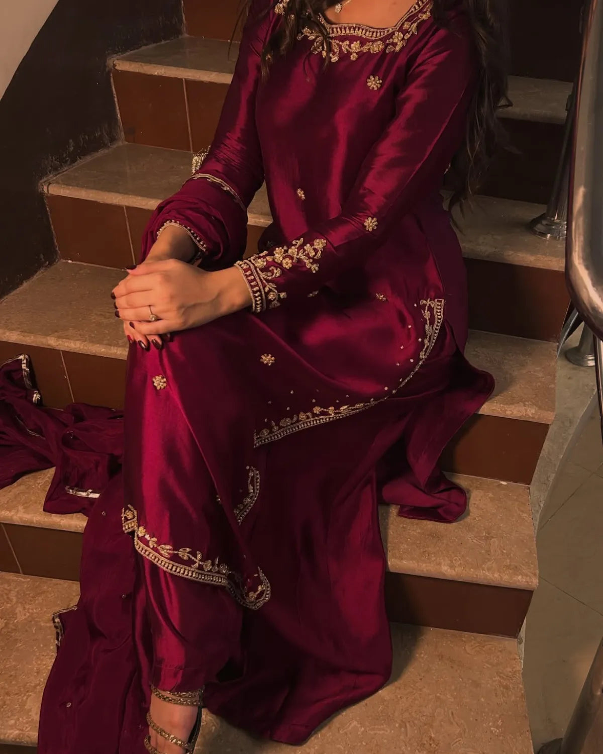 A woman seated on indoor stairs wearing a deep magenta satin traditional outfit with gold embroidery and a matching draped fabric on the floor.