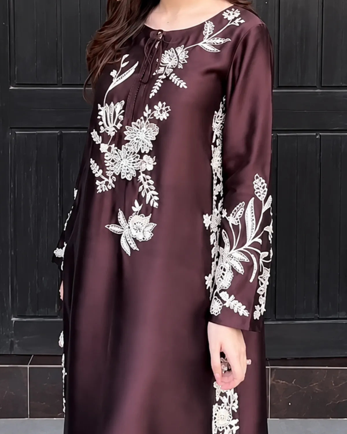 A woman wearing a long-sleeved, knee-length dark brown satin dress with intricate white floral embroidery, standing in front of a dark paneled door.