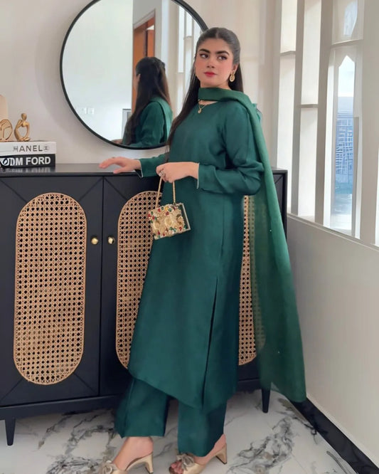 A woman models a dark green traditional outfit with matching dupatta, trousers, and a small floral handbag, standing in front of a black cabinet with woven panels and a large round mirror.