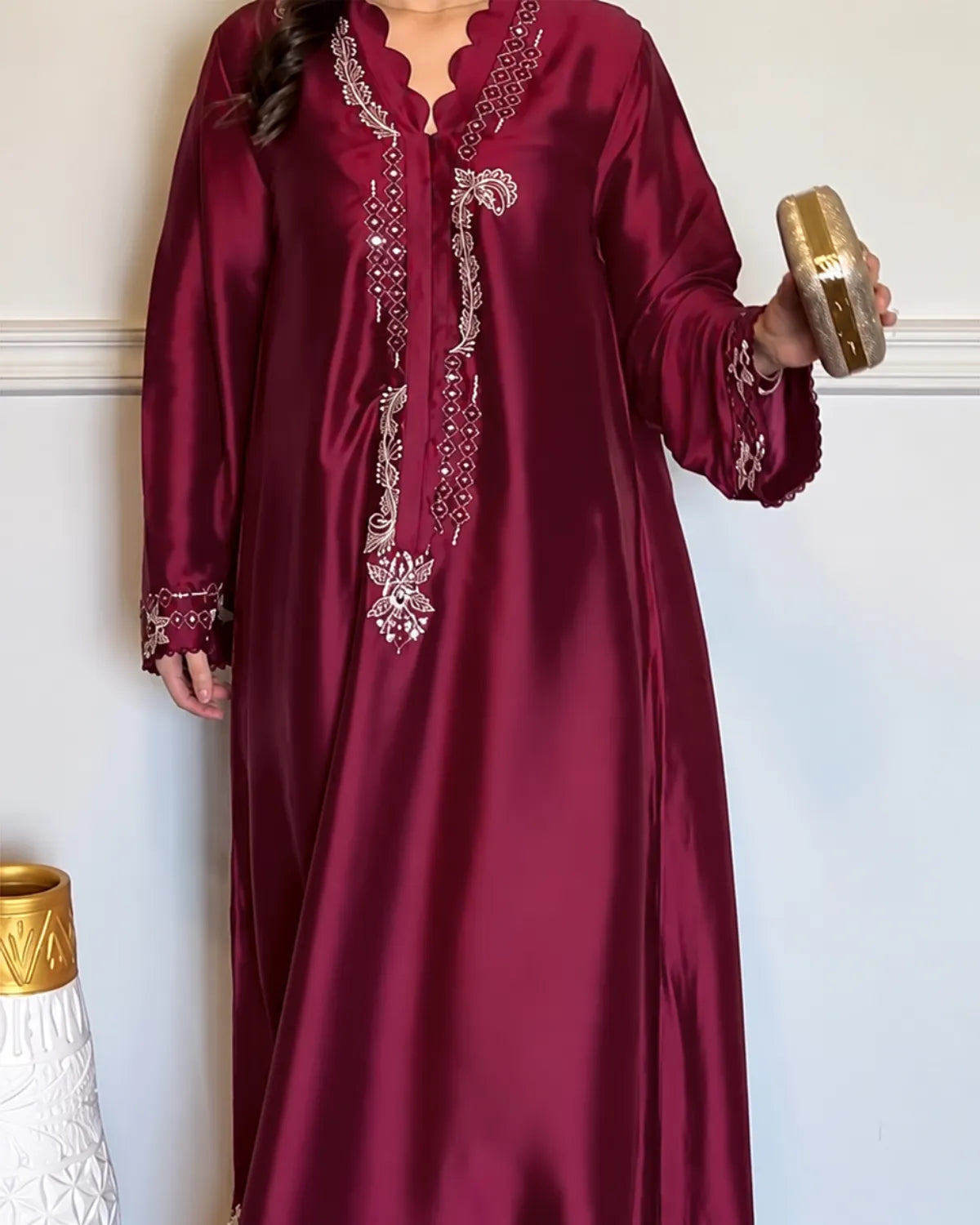 A woman wearing a long-sleeved, floor-length maroon satin abaya with intricate silver embroidery and beadwork on the front, neckline, and cuffs, holding a gold clutch.
