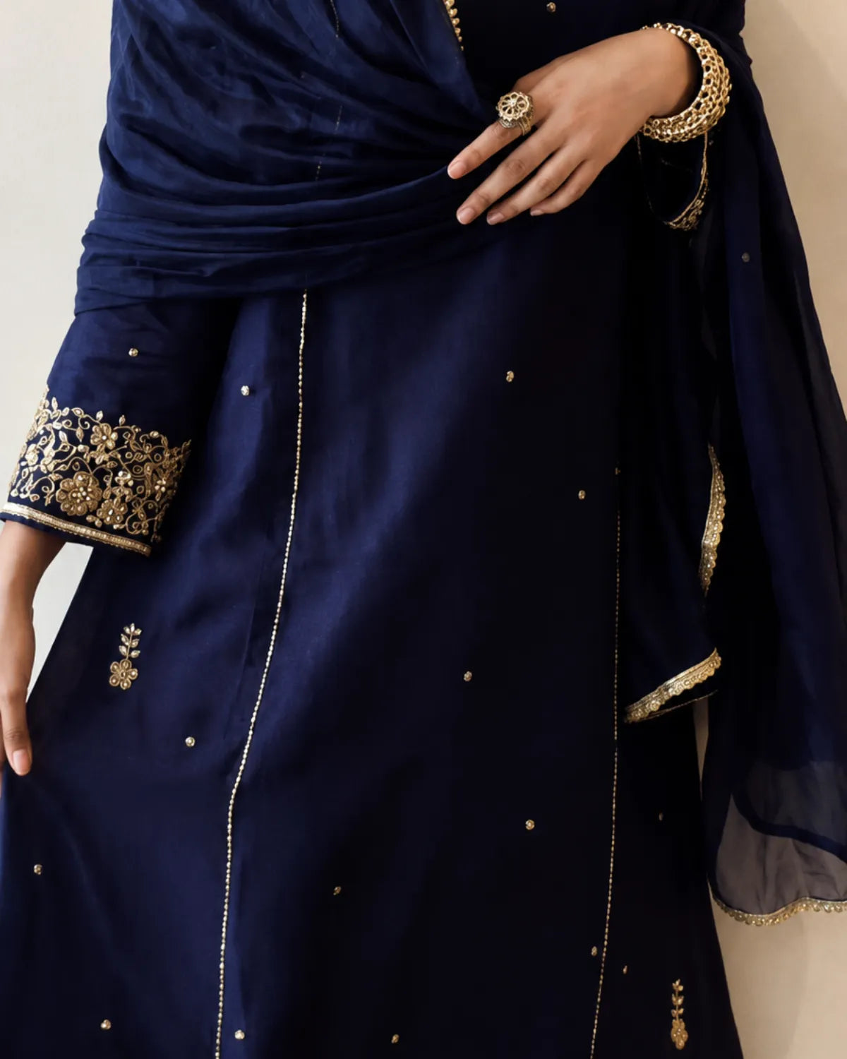 A close-up of a navy blue traditional outfit with gold embroidery on the sleeve cuffs, scattered gold dot embellishments, and a matching navy dupatta draped over the shoulder.