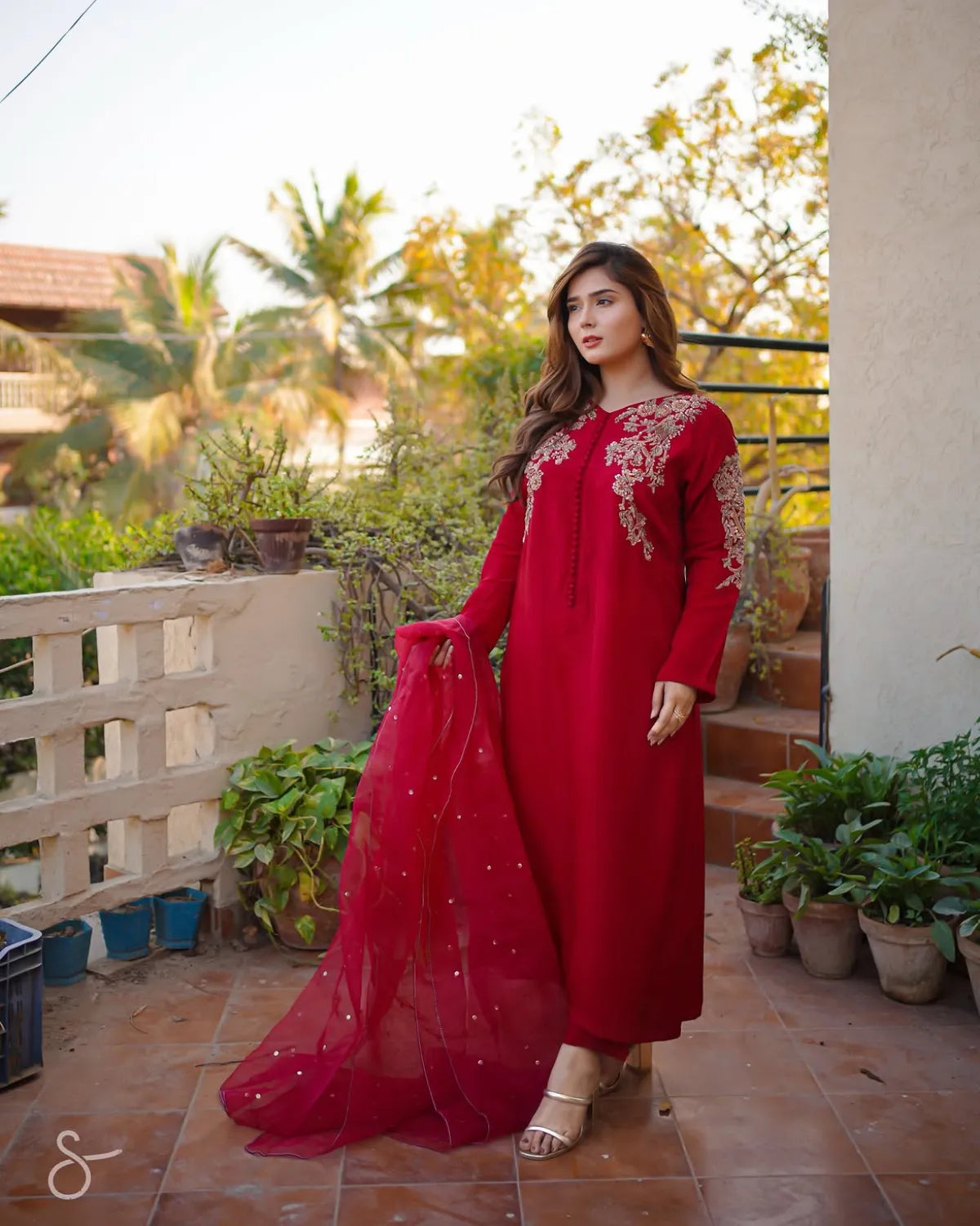 A woman wearing a long-sleeved red embroidered kurta with a matching sheer dupatta, standing on an outdoor balcony with plants and a tiled floor.