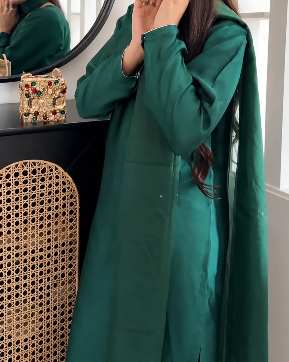 A woman wearing a long, flowing emerald green coat or dress, standing indoors near a black vanity table with a round mirror and a woven cane chair. The product appears to be the green outerwear or dress being modeled.