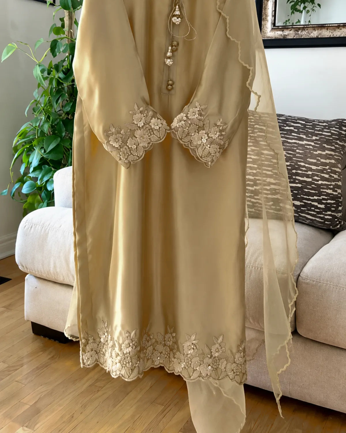 A beige satin kurta with matching sheer dupatta and floral lace embroidery, shown hanging in a living room setting.