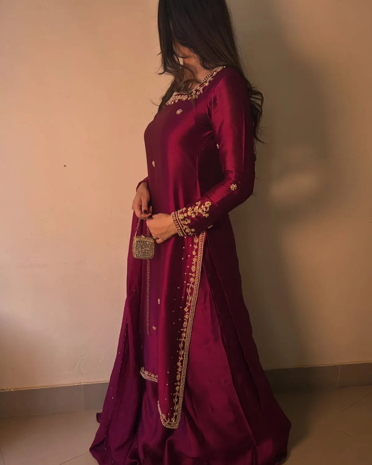 A woman wearing a deep magenta-purple traditional dress with gold embroidery, holding a small metallic clutch bag against a plain beige wall.