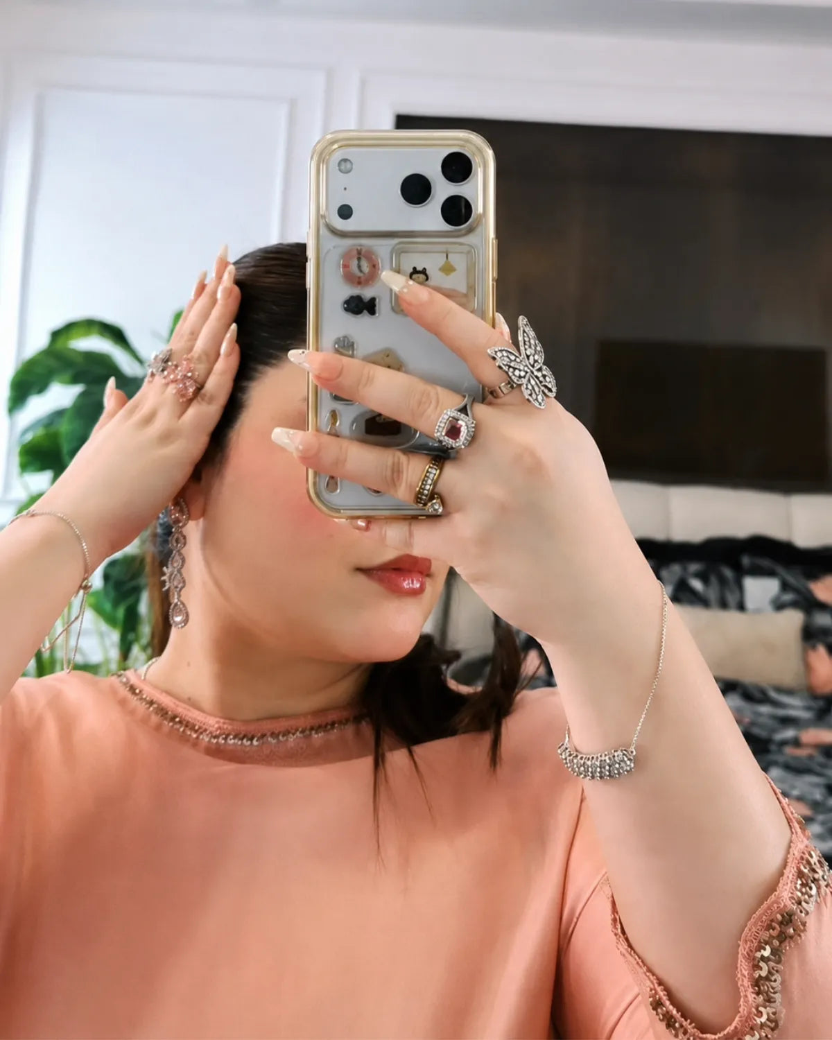 A woman taking a mirror selfie in a bedroom, showcasing a collection of silver-toned jewelry including rings, a bracelet, and earrings. She is wearing a peach-colored top and holding a phone with a decorative case.