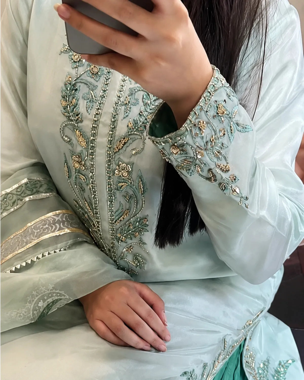 A close-up of a person wearing a light sage green traditional outfit with intricate gold and green embroidery on the chest and sleeve cuffs, holding a phone to take a mirror selfie.