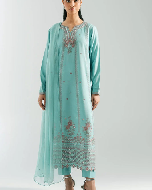 A woman modeling a mint green traditional kurta set with matching pants and a sheer dupatta, featuring detailed floral embroidery and metallic embellishments, set against a plain light gray background.