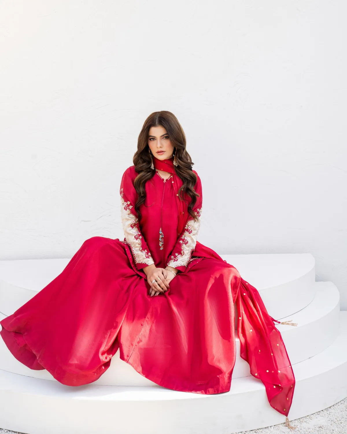 A woman wearing a bright red traditional outfit with white embroidered sleeves sits on white tiered steps against a white wall.