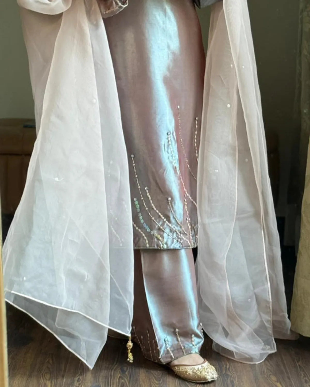 A person wearing a shimmering metallic taupe-pink traditional outfit with wide flared pants, embellished with vertical beadwork, and draped with a sheer white tulle overlay. Gold flat shoes are visible at the bottom.