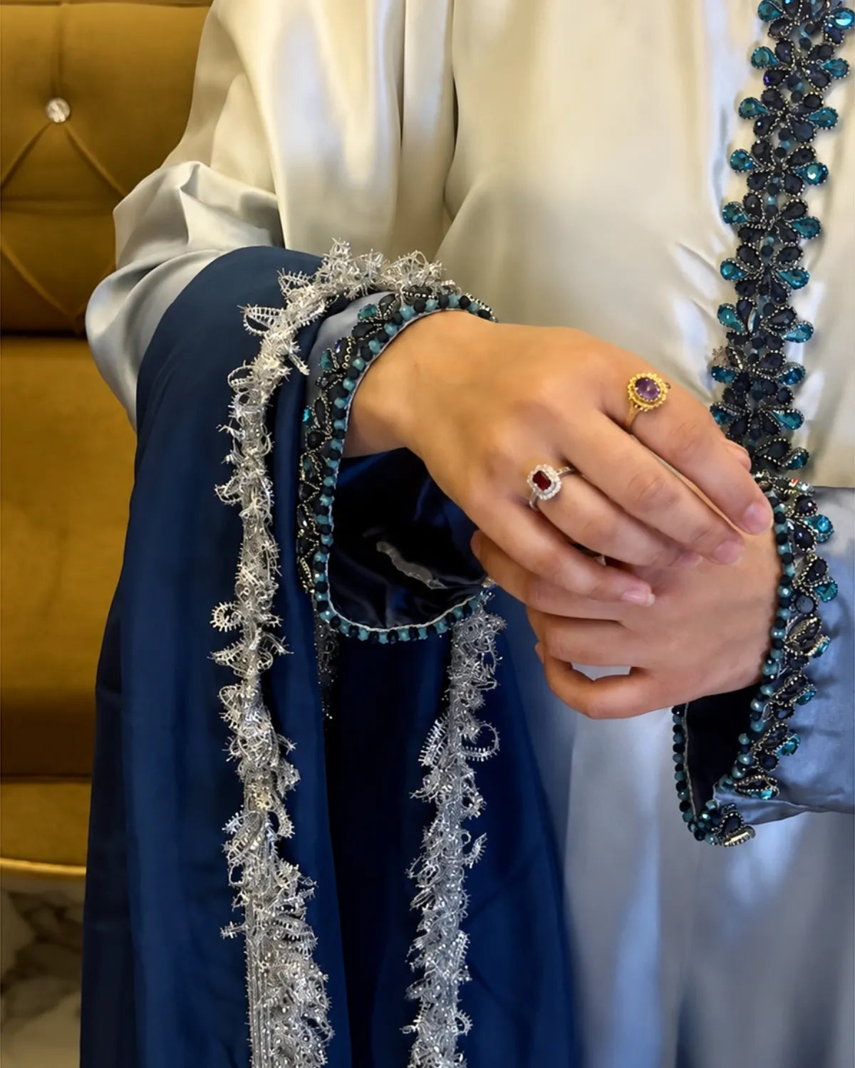 A close-up of a person wearing traditional South Asian attire, featuring a deep navy blue kurta with ornate silver and teal embroidery, paired with a white abaya. The person is shown from the chest down, with hands clasped together displaying two rings.