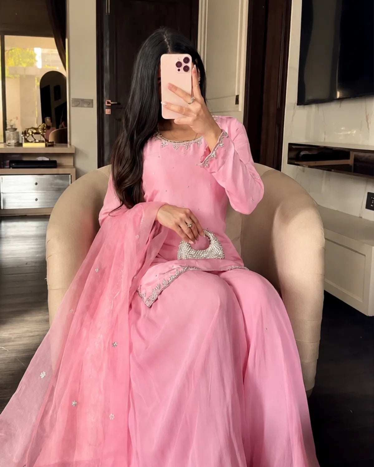 A woman takes a mirror selfie wearing a pink traditional long dress with silver embellishments, seated in a beige armchair inside a modern home interior.