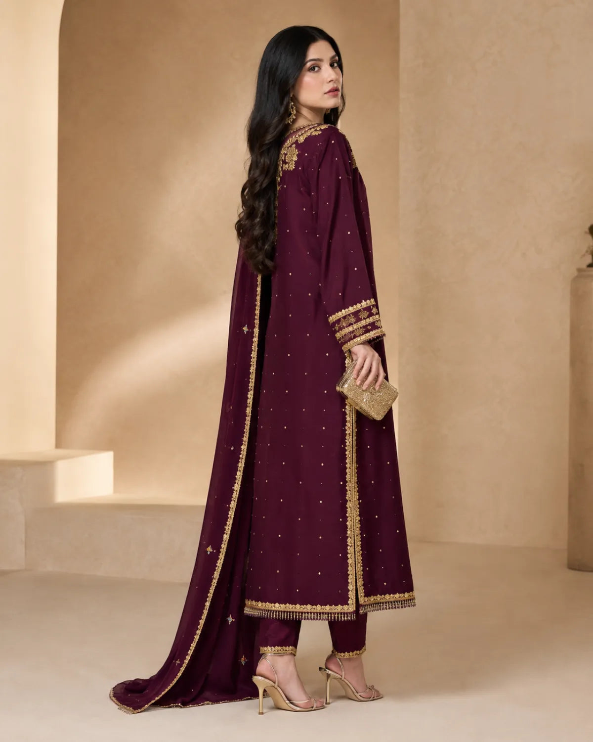 A woman modeling a deep maroon traditional outfit with gold embroidery and a matching dupatta, standing in a warm beige architectural setting.