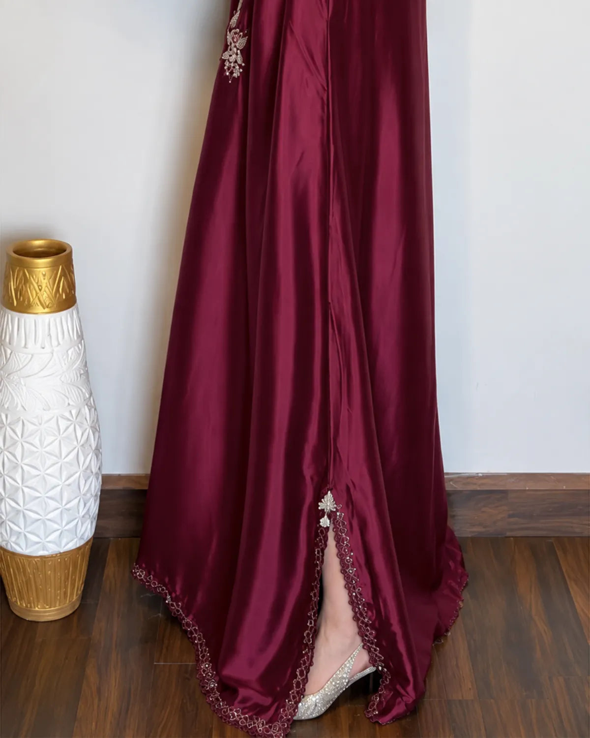 A floor-length maroon satin gown with a high slit, decorated with silver floral embroidery near the waist and a matching embroidered hem, shown on a model standing on a wooden floor next to a white and gold vase.