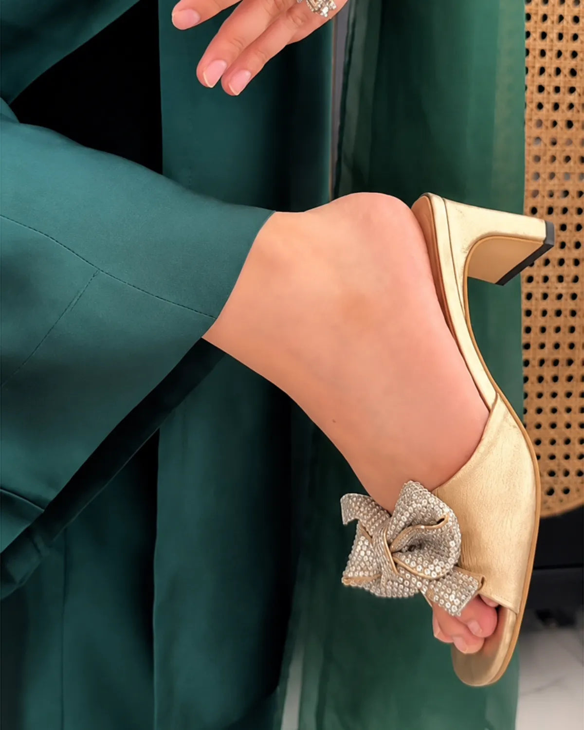 A close-up of a person wearing a metallic gold mule sandal with a large rhinestone-embellished bow on the side, paired with a dark green outfit.