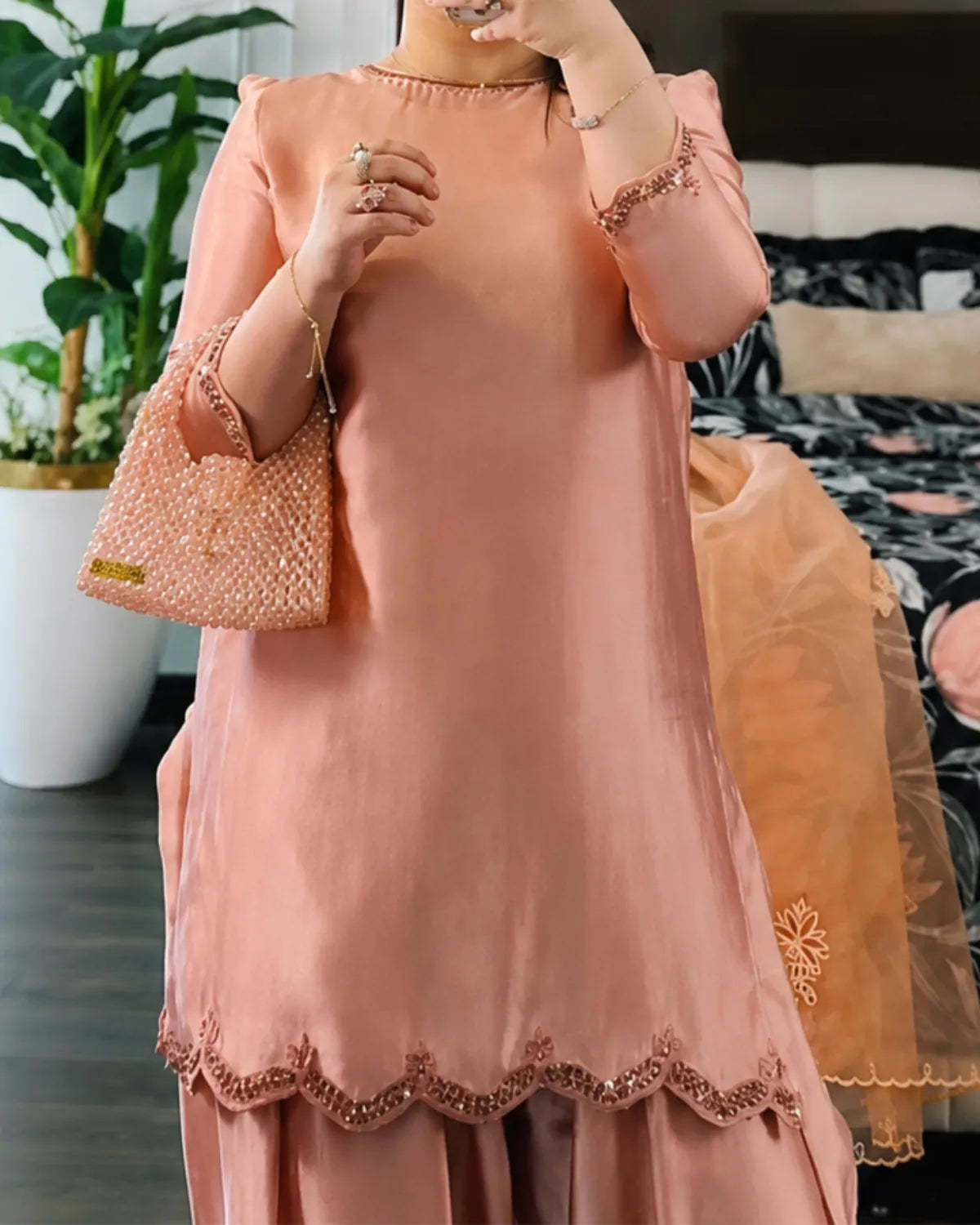 A woman wearing a peach-pink traditional kurta set with matching pants, holding a textured peach-pink handbag. The kurta features a scalloped embroidered hem and lace-trimmed sleeves, shown in a bedroom setting.