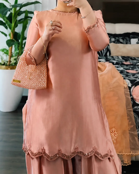 A woman wearing a peach-pink traditional kurta set with matching pants, holding a textured peach-pink handbag. The kurta features a scalloped embroidered hem and lace-trimmed sleeves, shown in a bedroom setting.