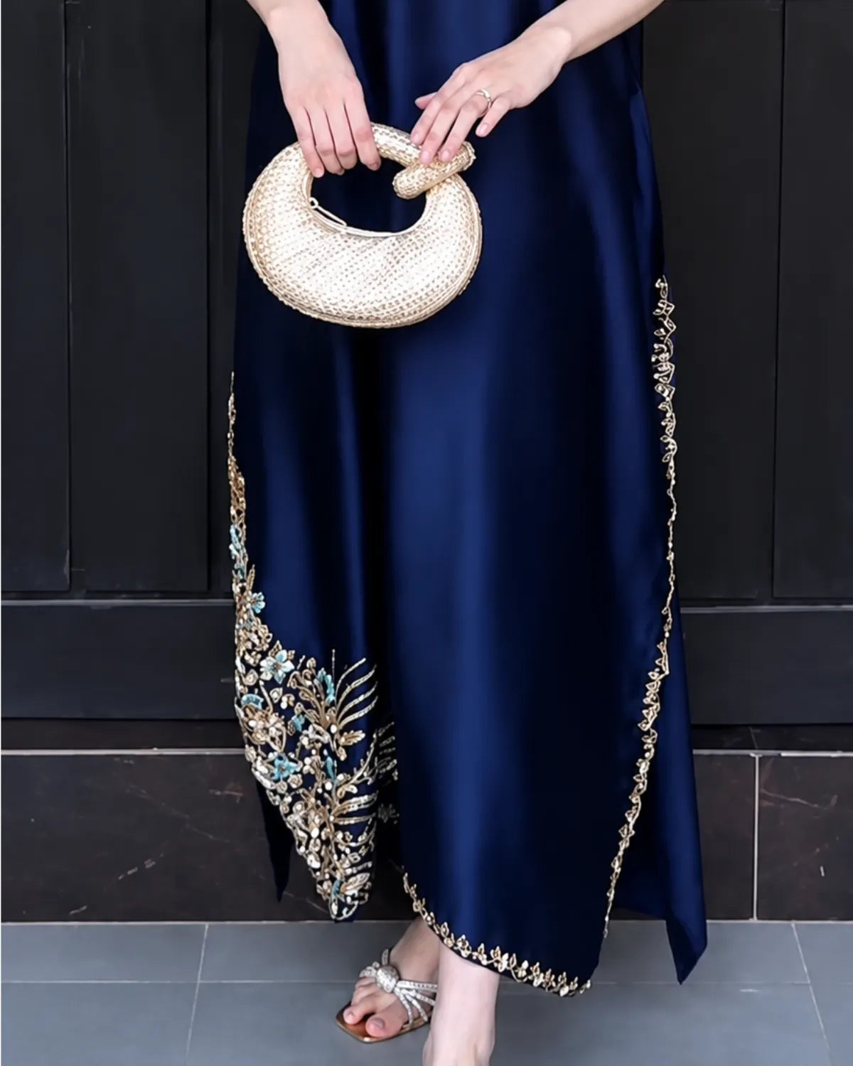 A navy blue satin maxi dress with intricate gold, silver, and blue floral embroidery and gold border detailing, shown on a model holding a woven straw handbag.